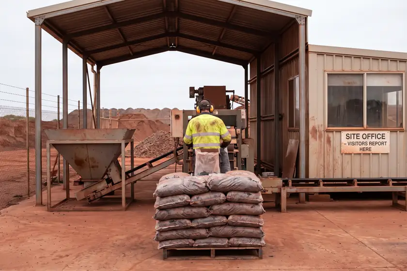 A worker in hi-vis and ear muffs stacking filled bags onto a pallet under an open-sided bagging shed, hopper and conveyor behind.