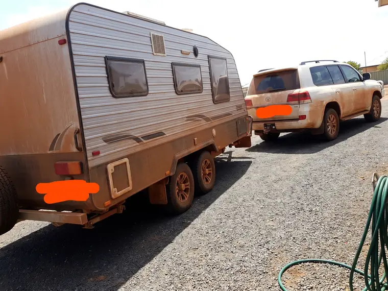 Customer photo of a tandem caravan on a gravel driveway, coated in red dust along its full flank, with an equally dusty LandCruiser wagon hitched to it. The number plate has been scribbled out.